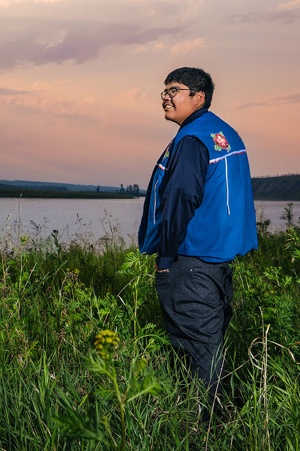 Samuel Metacat-Yah standing in tall grass in front of a river and a peachy sunset.
