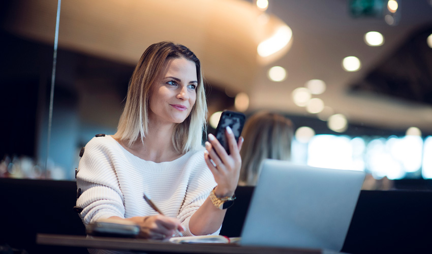 A business woman using holding a smartphone; sitting in front of a laptop.