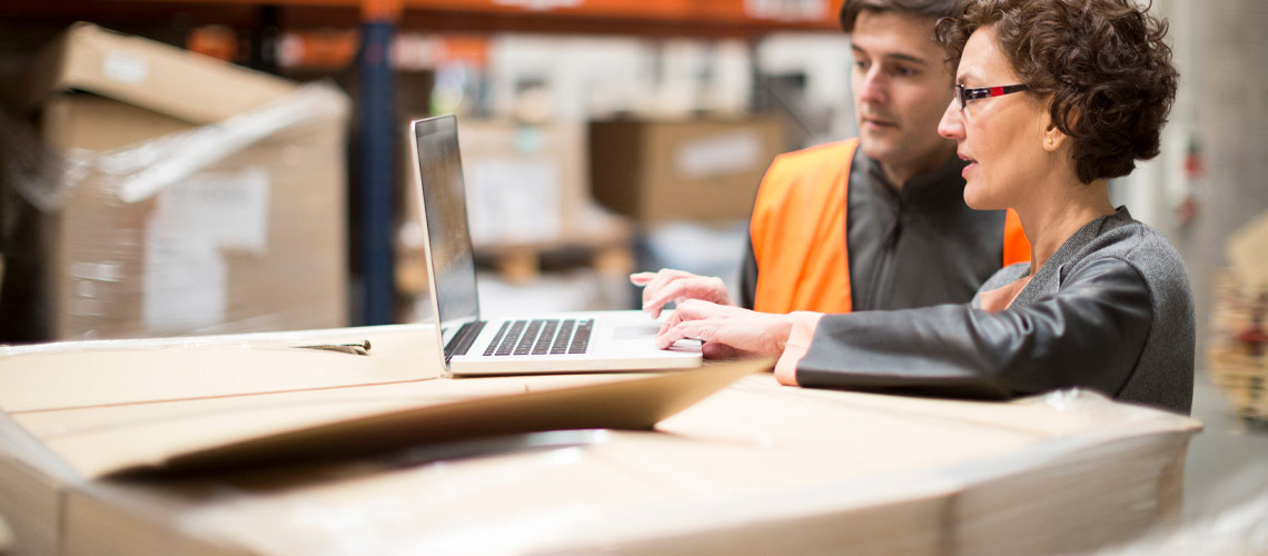 Two warehouse workers having a meeting, one is working on a laptop.