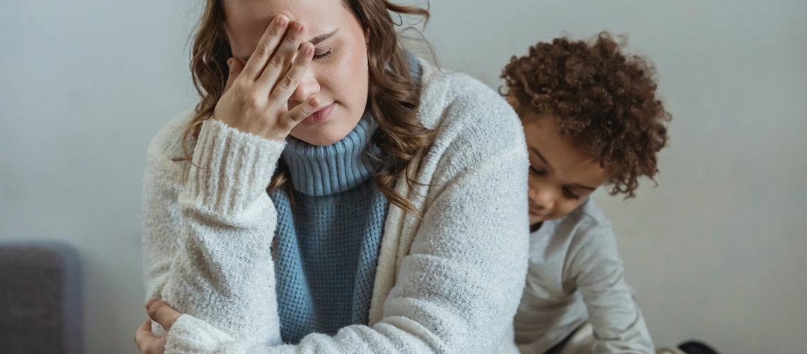 A mom sitting on couch with hand on face; her child is playing in the background.