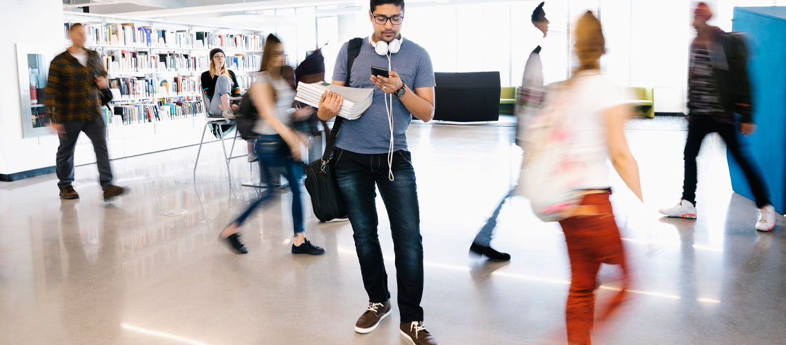 A student standing in the MRU Library looking at his iPhone while students walk in the background.