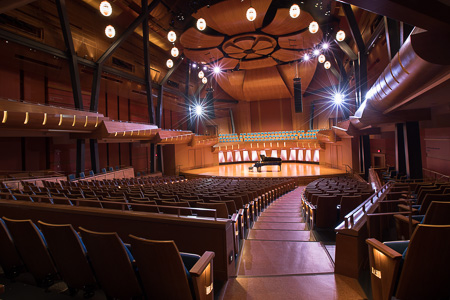 The seating and stage of a concert hall. Everything is made out of warm wood and a series of curved wood panels hangs above the stage.