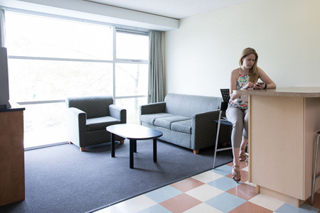 A woman sitting at a kitchen counter in front of a simple living room seating area.