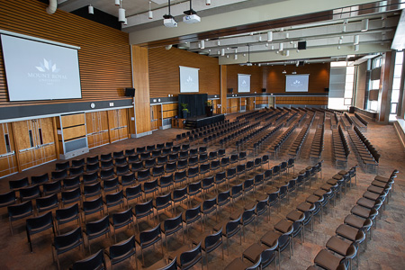 The hall of a conference centre with rows of chairs, a temporary stage and large screens.