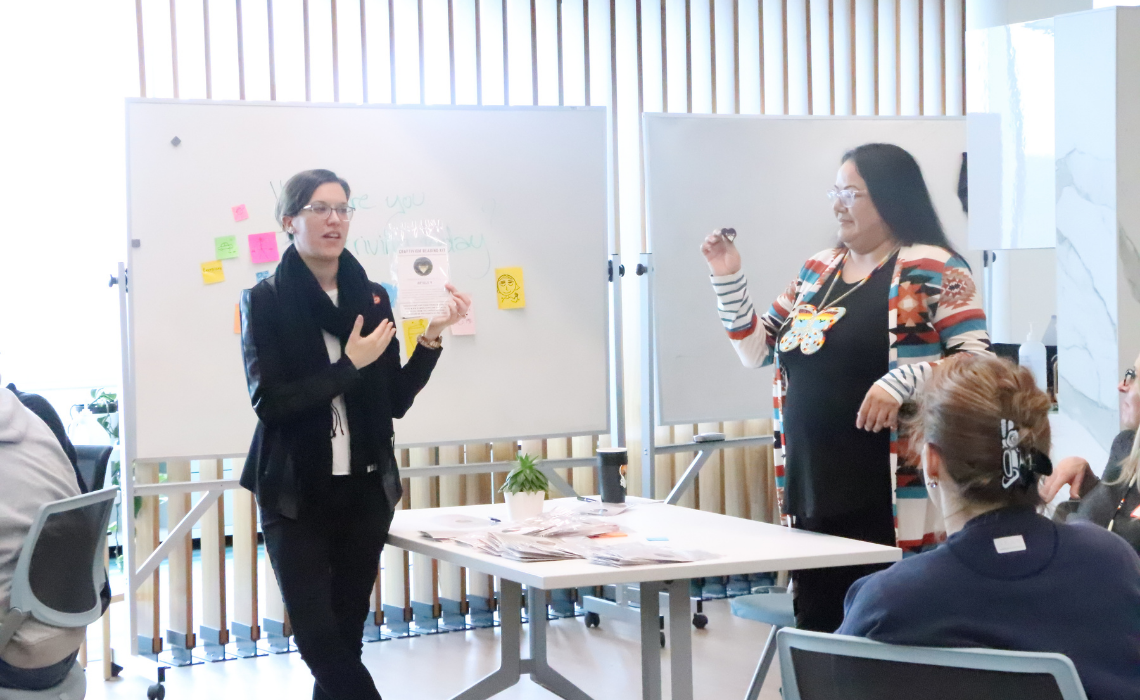 Two people leading a discussion at the front of a room, with whiteboards, sticky notes, and seated participants.