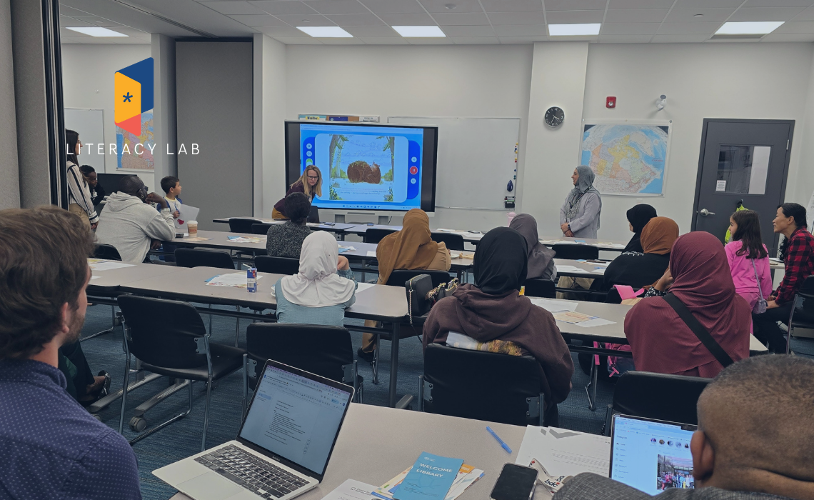Teacher leads a literacy workshop while attendees take notes on laptops; classroom setting with maps and handouts visible.
