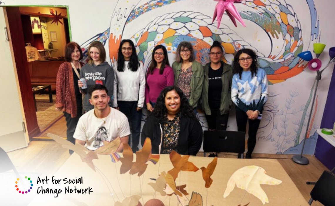 A group of nine people is gathered indoors in front of a colorful mural featuring a large, abstract, snake-like design with scales in various colors such as orange, blue, and green.    The individuals are standing and sitting around a table that has a decorative mobile with bird shapes hanging above it. The room has wooden flooring and a cozy atmosphere with a lamp featuring multiple colored shades on the right side.  In the bottom left corner, there is a logo with a circular, multicolored dotted design and the text "Art for Social Change Network," indicating the context or organization related to the group.