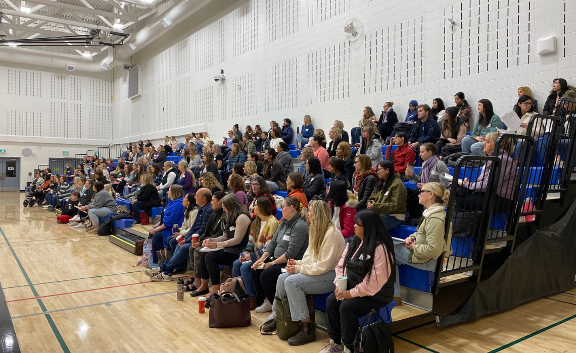 A large group of people seated on blue bleachers inside a gymnasium. The audience appears attentive, facing towards the left side of the image, suggesting they are watching or listening to a presentation or event. The gym has a high ceiling with visible lighting fixtures and ventilation ducts, and the floor is polished wood with sports court markings.