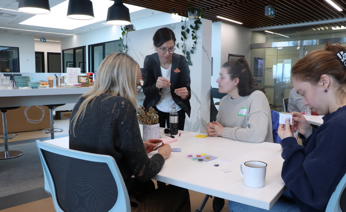 A group of people sitting together at a table in a bright, modern workspace, talking and working with small craft materials.