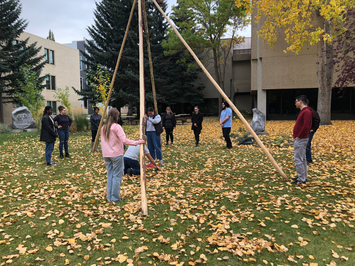 A group of five Catamount students on the MRU campus, building a tipi in a yard with the beautiful fall foliage as a backdrop.
