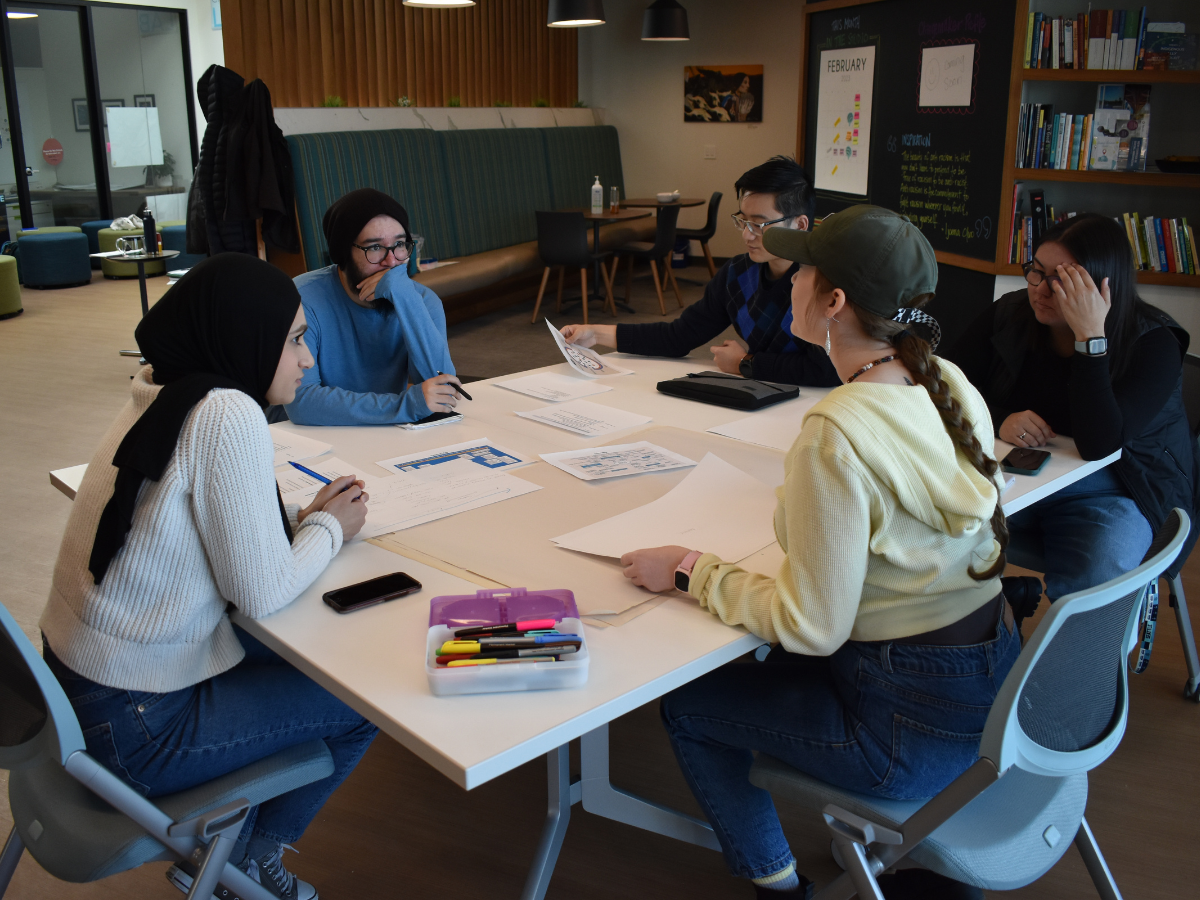 A group of 4 students at Mount Royal University are engaging in conversations around a square table in the Trico Changemakers Studio